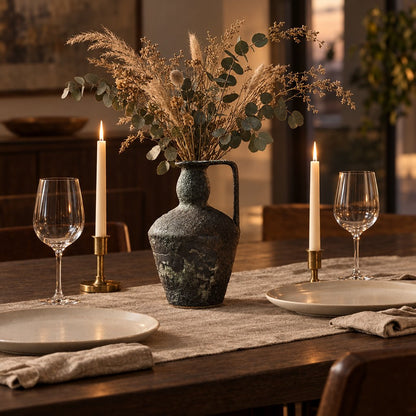 Dark charcoal-green rough-textured ceramic pitcher vase holding dried pampas, golden filler flowers and eucalyptus on a dining table with lit taper candles, wine glasses and cream plates