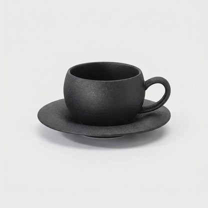 Matte charcoal-black stone-finish ceramic cup with a loop handle on a wide shallow saucer, shown at a three-quarter angle on a plain light gray background