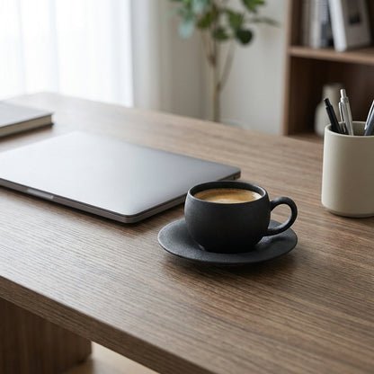 Matte charcoal-black stone-finish ceramic cup and saucer holding espresso on a wooden desk beside a closed laptop, a notebook and a pencil cup