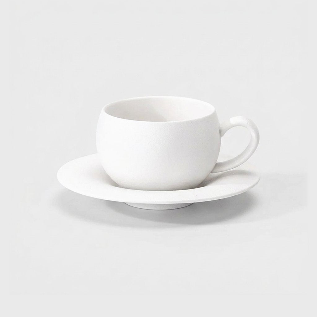 Matte white stone-finish ceramic cup with a loop handle on a wide shallow saucer, shown at a three-quarter angle on a plain light gray background