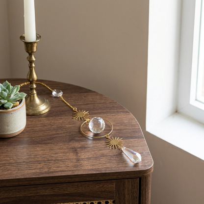 Decorative suncatcher crystal and brass chain on a wooden table with a candle and plant in the background.