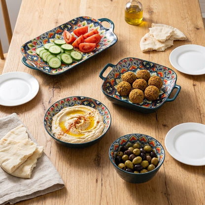Overhead view of four patterned ceramic serving pieces holding cucumber tomato salad, falafel, hummus and olives on an oak table with pita