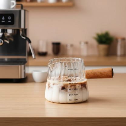 Glass milk frothing pitcher on kitchen counter beside espresso machine