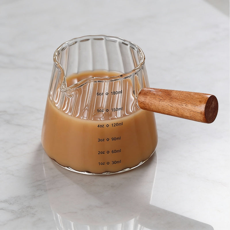 Glass measuring cup with wooden handle filled with a brown liquid on a marble surface