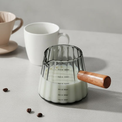 Glass milk pitcher with wooden handle on a light surface with coffee cups and beans.