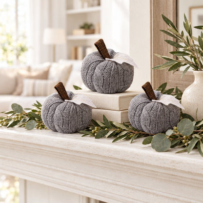Three grey cable-knit mini pumpkins on a white mantel with a eucalyptus garland, one raised on a stack of cream books
