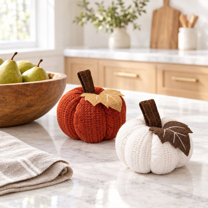 Orange and cream mini knit pumpkins on a white marble kitchen island beside a wooden bowl of green pears and a striped towel