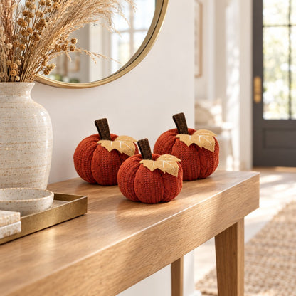 Three rust orange mini knit pumpkins on an entry console table under a round brass mirror with a speckled vase of dried grasses