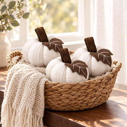 Three cream mini knit pumpkins with brown stems and leaves in a woven basket on a window ledge with a chunky knit throw