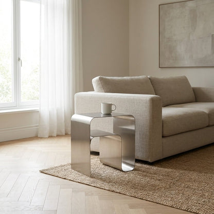 Brushed stainless steel arch side table at the arm of a beige sofa holding a white ceramic mug, with an empty lower shelf
