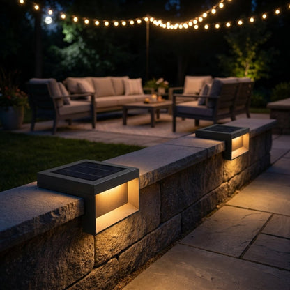 Two black solar deck lights glowing warm on a low stone wall cap beside a patio with a gray sofa, armchairs and string lights