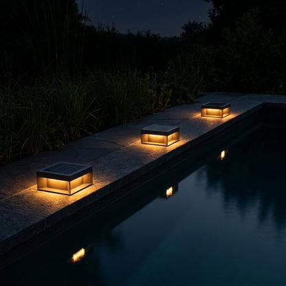 Three black solar deck lights glowing warm amber on the gray stone edge of a pool at night with tall grasses behind