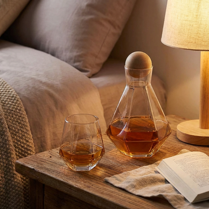Glass decanter and glass with amber liquid on a wooden table next to a lamp and book.