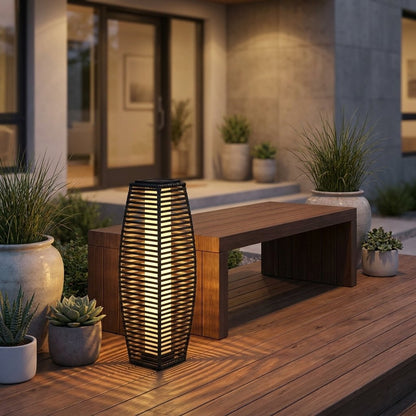 Lit outdoor solar floor lantern with woven rattan panels on a wood deck beside a bench, surrounded by potted succulents and ornamental grasses at dusk