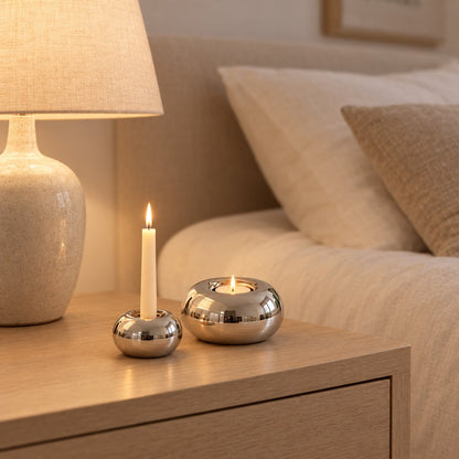 Small polished silver candleholder with a lit white taper candle and a larger one with a lit tealight on a light wood bedside table beside a cream speckled lamp with a linen shade
