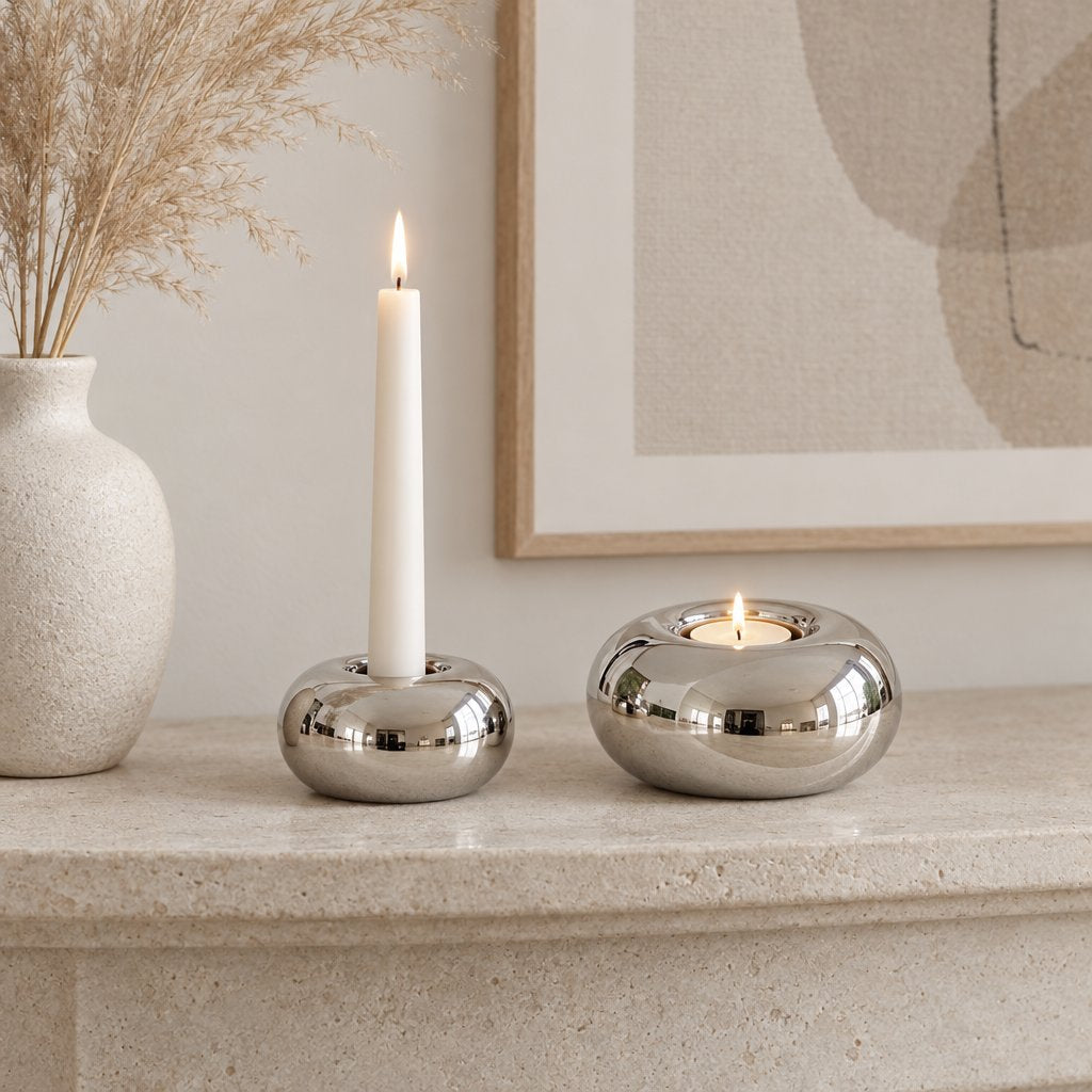 Small polished silver candleholder with a lit white taper candle and a larger one with a lit tealight on a pale stone mantel beside a speckled vase of pampas grass and a framed abstract print