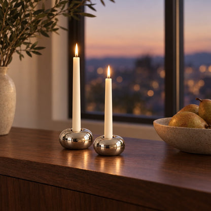 Two small polished silver rounded candleholders each with a lit cream taper candle on a dark walnut console beside a speckled cream vase of olive sprigs, with a twilight city window behind