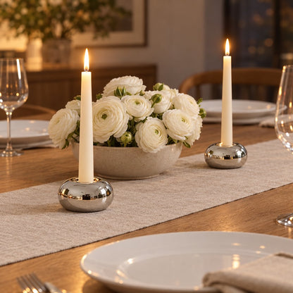 Two small polished silver rounded candleholders with lit cream taper candles on a beige runner on a wooden dining table with a speckled bowl of white ranunculus, wine glasses and plates