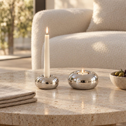 Small polished silver candleholder with a lit white taper candle and a larger one with a lit tealight on a round pitted travertine coffee table beside a stone bowl of olives with rosemary and a white boucle armchair