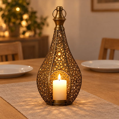Bronze cutwork metal lantern with a lit pillar candle on a linen runner on a wood dining table, with plates and chairs in the background