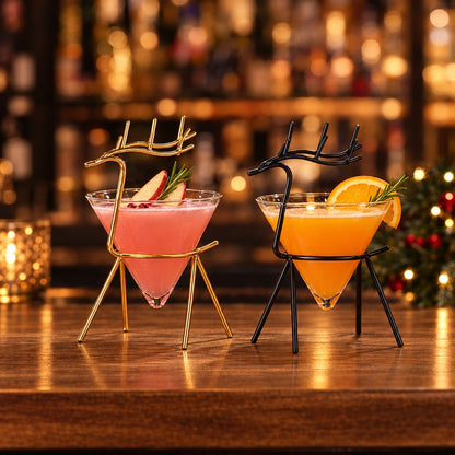 Gold and black reindeer stand cocktail glasses on a wooden bar counter, one holding a pink drink with apple slices and rosemary and one holding an orange drink with an orange slice, with bottles and Christmas lights behind