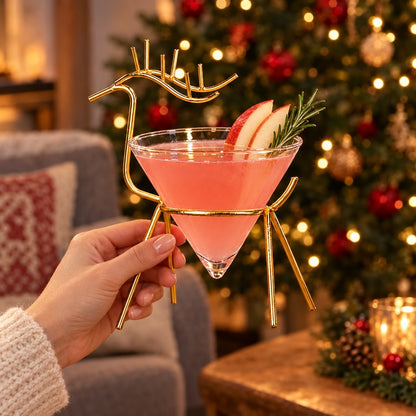 Hand holding a gold reindeer stand cone cocktail glass with a pink drink, apple slices and rosemary in front of a decorated Christmas tree