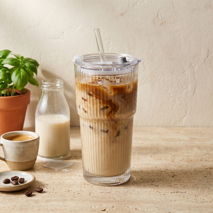 Ribbed glass tumbler with iced coffee on white table beside plant