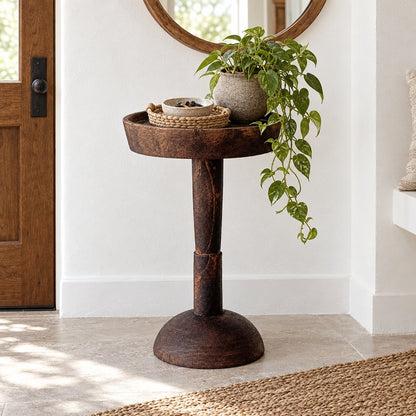 Large dark wood round pedestal side table in an entryway holding a trailing green plant, a woven basket and a small bowl beneath a round mirror beside a wooden door