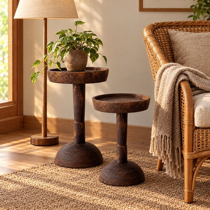 Two dark wood round pedestal side tables in different heights beside a rattan armchair with a knit throw and a floor lamp, the taller one holding a potted plant