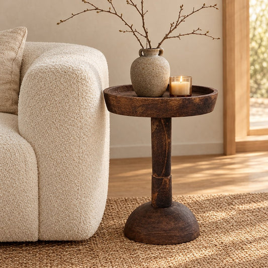 Dark wood round pedestal side table with a shallow raised rim beside a boucle sofa holding a grey ceramic vase with bare branches and a lit candle