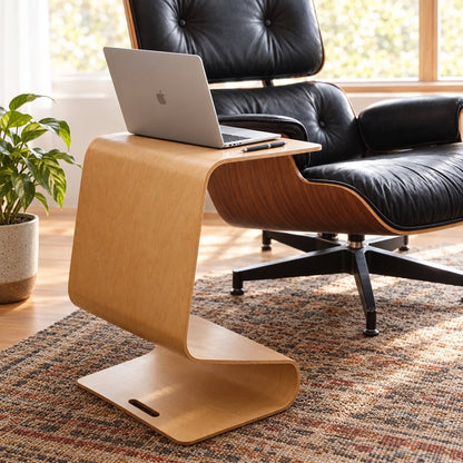 Light wood curved side table with a slot at one end, holding a laptop in front of a black lounge chair on a patterned rug, with a potted plant at left near a window