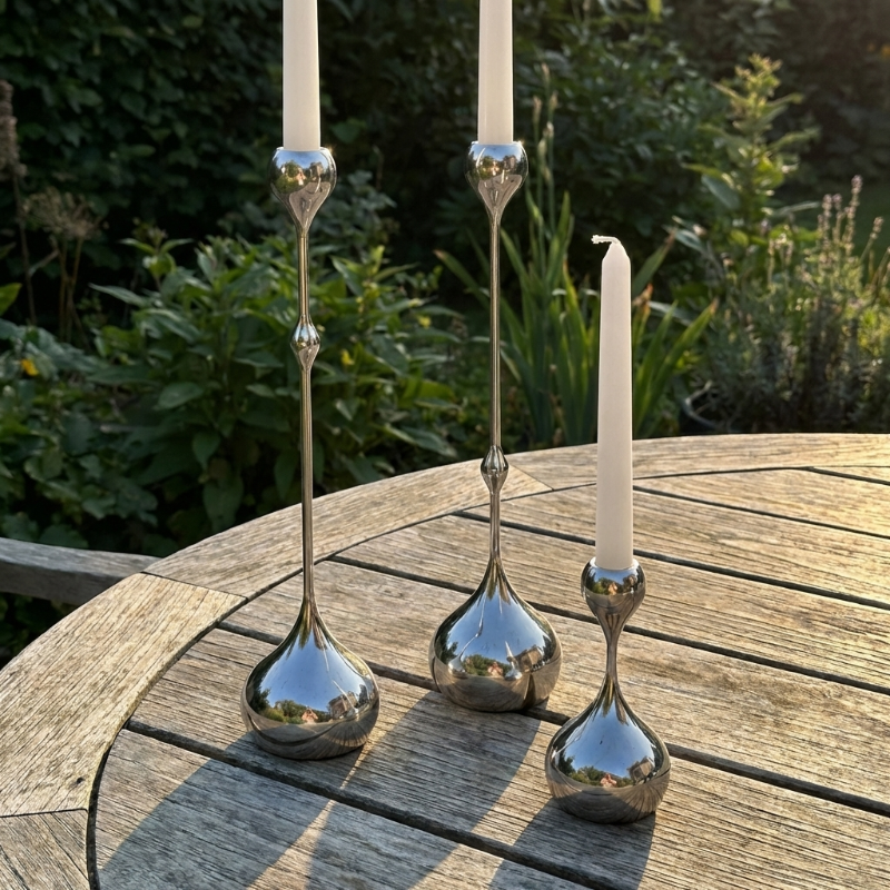 Three silver candle holders with candles on a wooden table outdoors.