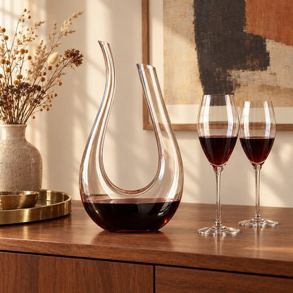 Clear U shaped glass wine decanter holding red wine on a wooden sideboard beside two glasses of red wine, a brass tray and a vase of dried stems