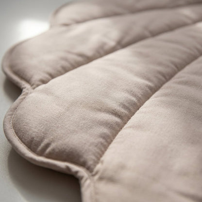 A close-up of a soft, quilted cushion or pad in a light taupe-beige fabric, showing the padded channels and scalloped edge against a neutral light background.