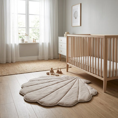 A scalloped shell-shaped quilted baby play mat in a light taupe-beige color, styled on a light wood floor in a bright, modern nursery next to a wooden crib.