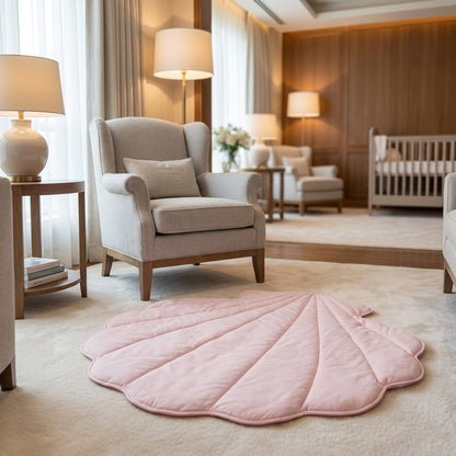 A pink shell-shaped quilted baby play mat on a beige carpet in a softly lit nursery with a light gray armchair, side table, lamps, and a crib in the background.