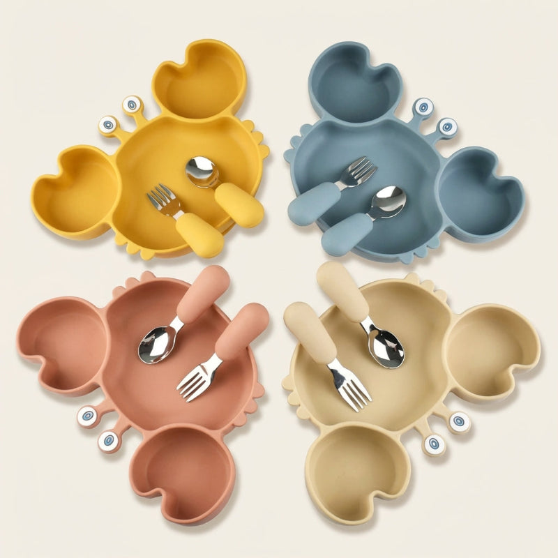 Four crab-shaped silicone divided plates in yellow, blue, pink, and beige, each with a matching fork and spoon set, arranged in a square on a white background.
