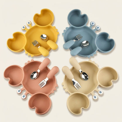 Four crab-shaped silicone divided plates in yellow, blue, pink, and beige, each with a matching fork and spoon set, arranged in a square on a white background.