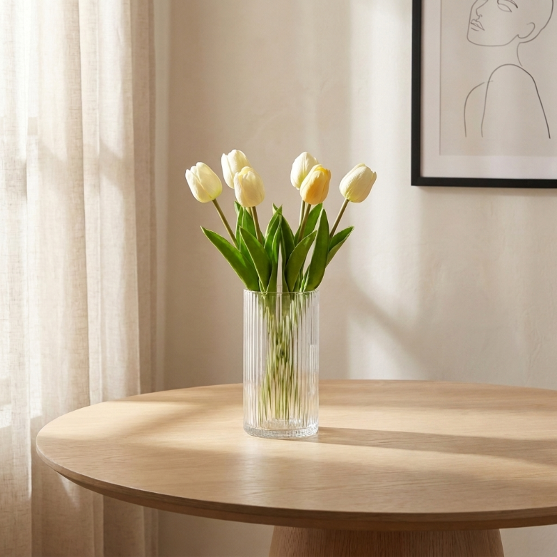 Clear vase with white and yellow tulips on a wooden table against a neutral wall with a framed picture.