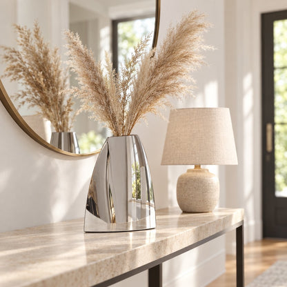 Large mirror silver arched vase with pampas on a travertine top console beside a round brass framed mirror and a beige textured table lamp