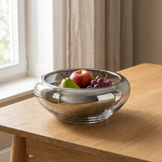 Mirrored silver glass bowl on a wooden dining table filled with an apple, pear, and grapes, in front of a sunlit window