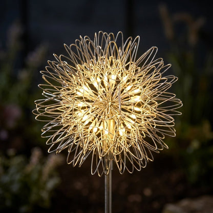 Close-up of a dandelion-shaped wire solar light head glowing warm white against a dark blurred garden