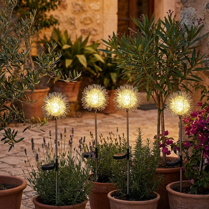 Four dandelion-shaped solar lights glowing among terracotta pots of lavender and olive trees on a stone courtyard at dusk