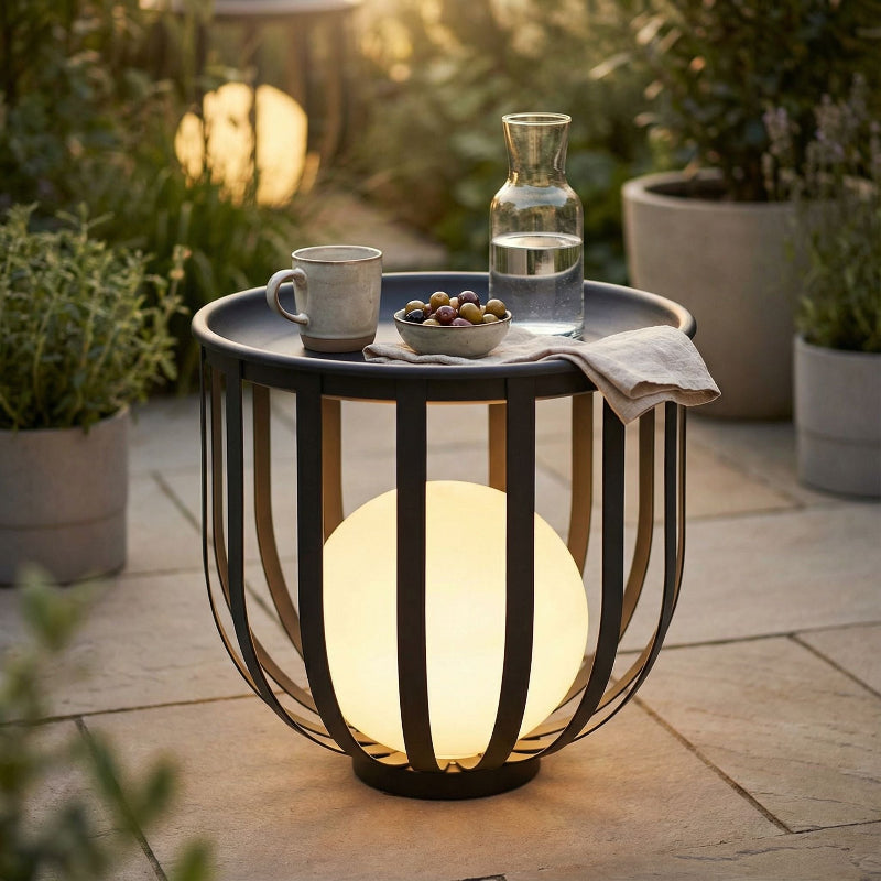 Outdoor setting with a small table holding a cup, bowl of snacks, and carafe of water, illuminated by a spherical lamp.