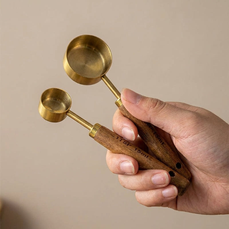 Hand holding two brass measuring spoons with wooden handles against a beige background