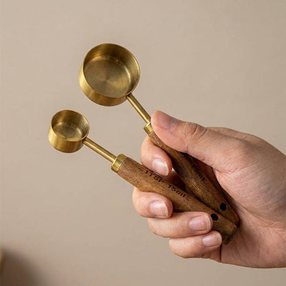 Hand holding two brass measuring spoons with wooden handles against a beige background