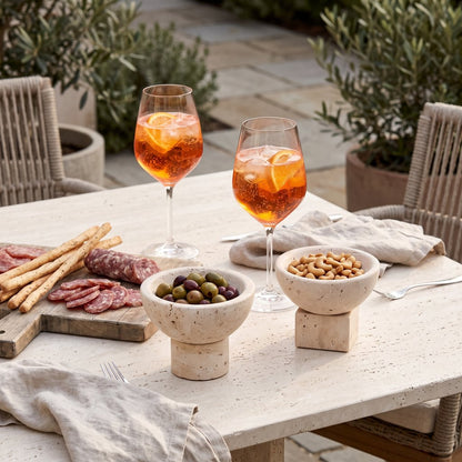 Two pale stone pedestal bowls with olives and nuts on a white outdoor table, with two orange spritz cocktails, sliced cured meats and breadsticks on a board
