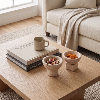 Two small pale stone pedestal bowls holding fruit on a wood coffee table with a stack of books and a mug, in front of a cream sofa with a throw