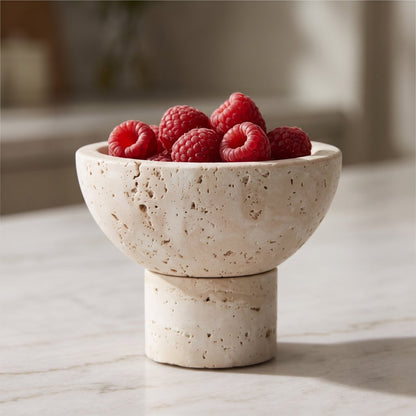 Close-up of a round pale stone pedestal bowl filled with raspberries on a marble counter