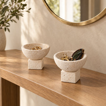 Two pale stone pedestal bowls with square bases holding small items on a wood console table beside a gold-framed round mirror and a plant stem